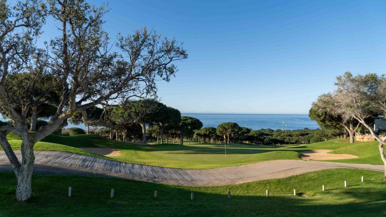 Blick auf einen Golfplatz mit gelber Fahne, im Vordergrund führt ein Weg entlang, im Hintergrund mehrere Bäume und Meer