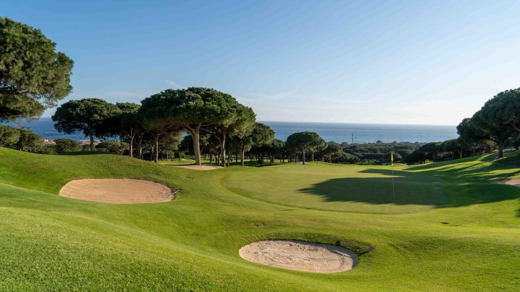 Blick auf Golfplatz Cabopino Marbella, mit gelber Fahne, daneben Bunker und Bäume, im Hintergrund Meer
