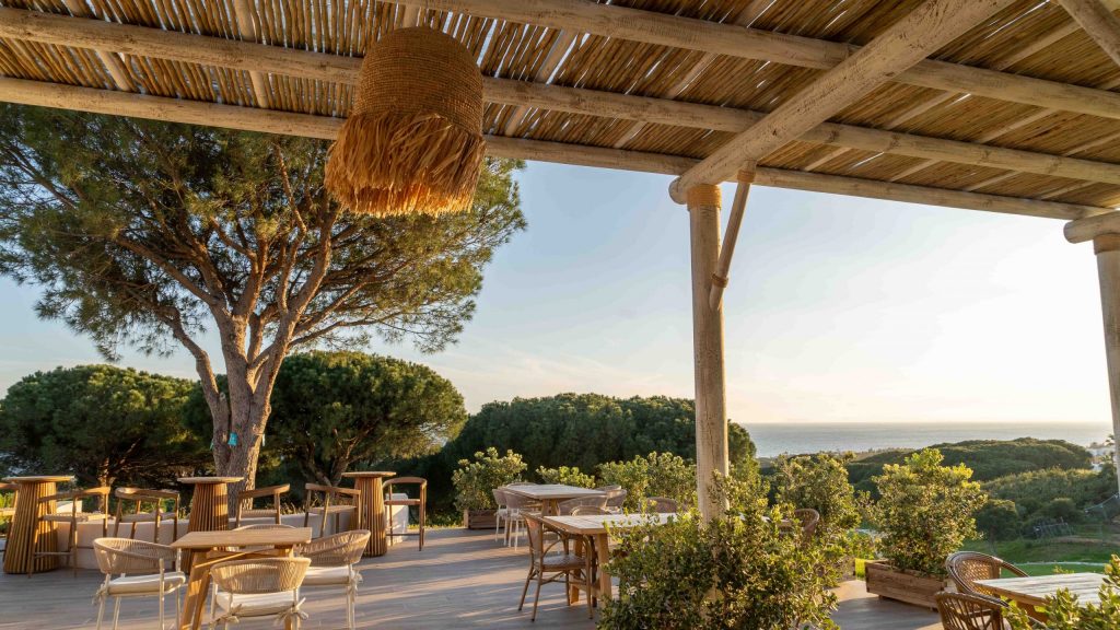 Außenbereich vom Restaurant des Cabopino Golf Marbella, Terrasse mit Stühlen und Tischen, daneben Bäume, im Hintergrund Meer