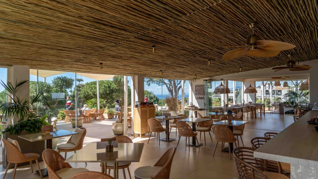 Restaurant des Cabopino Golf Marbella mit Bar, Tischen und Stühlen, Blick nach draußen auf die Terrasse, mit Bäumen und Meer im Hintergrund
