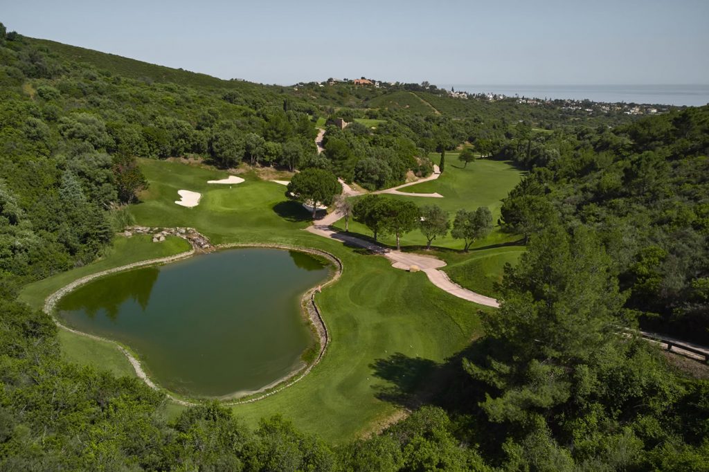 Blick auf die Golfanlage im Marbella Golf & Country Club mit einem kleinen See im Vordergrund, Platz wird von Wald umrandet, im Hintergrund grüne, hügelige Landschaft mit Häusern und Meer